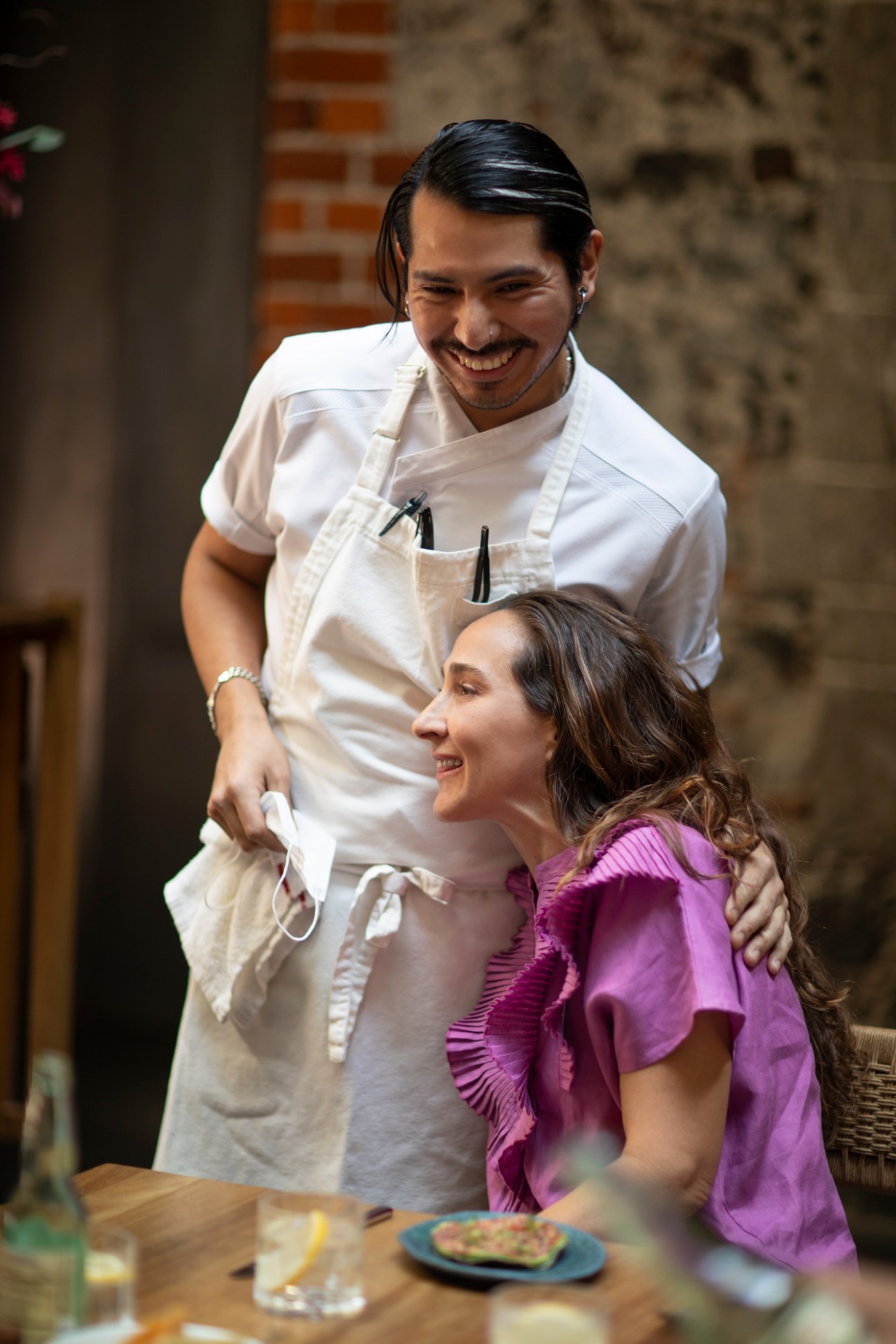 Chef in white uniform embracing woman in purple dress at restaurant table