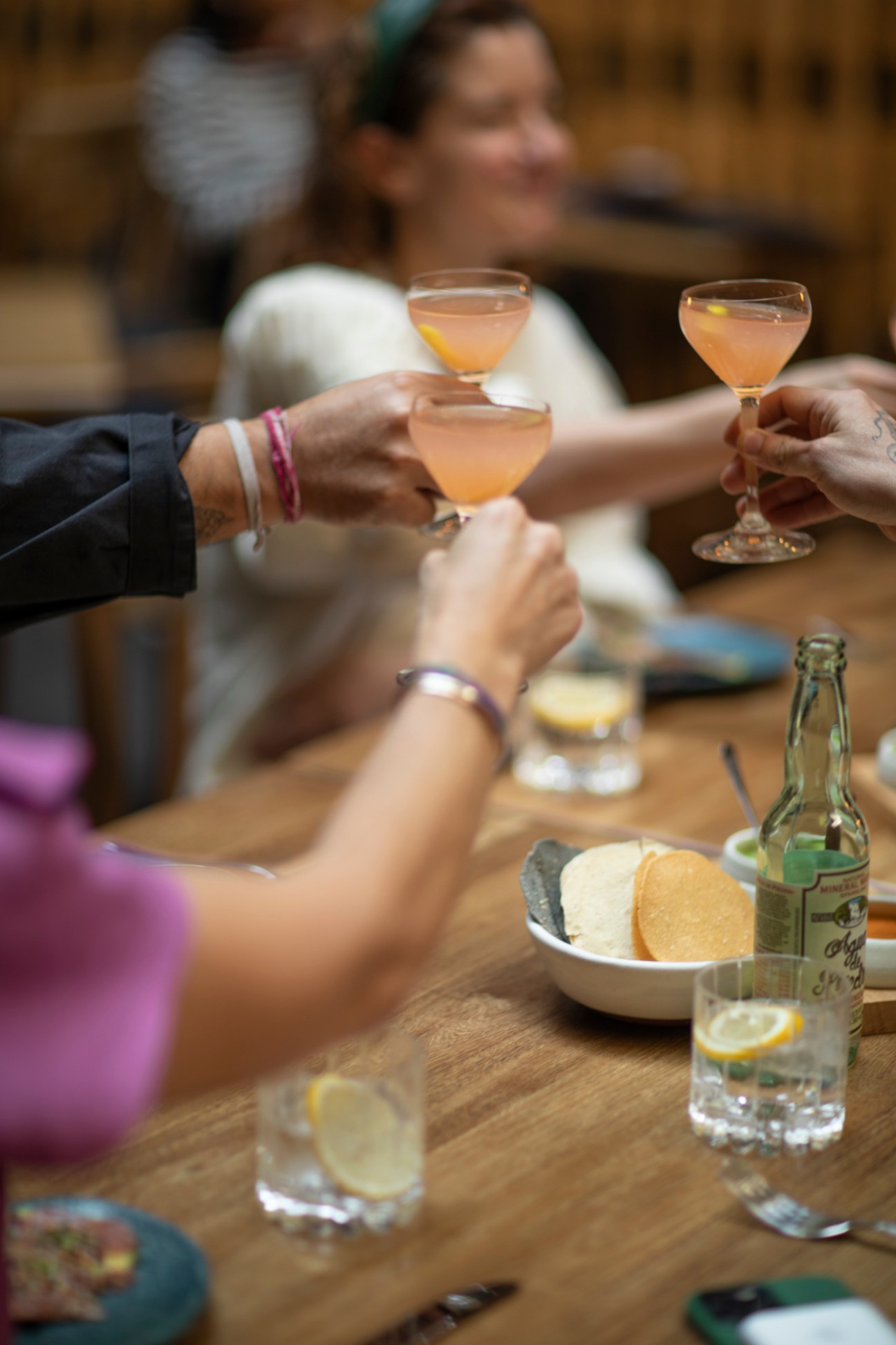 Hands raising cocktail glasses in a toast over a wooden table with snacks and bottles