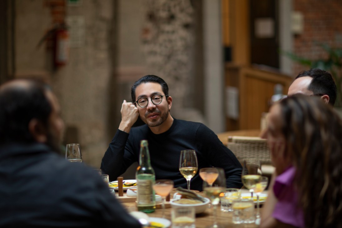 Man with glasses and dark sweater sitting at restaurant table with wine glasses and food during dinner conversation