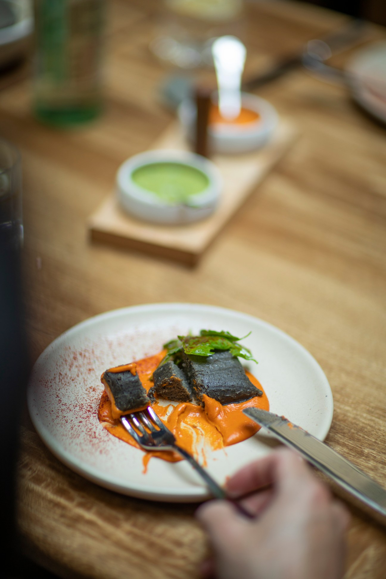 Plated dish with orange sauce, dark seaweed sheets, and green garnish being cut with fork and knife on wooden table