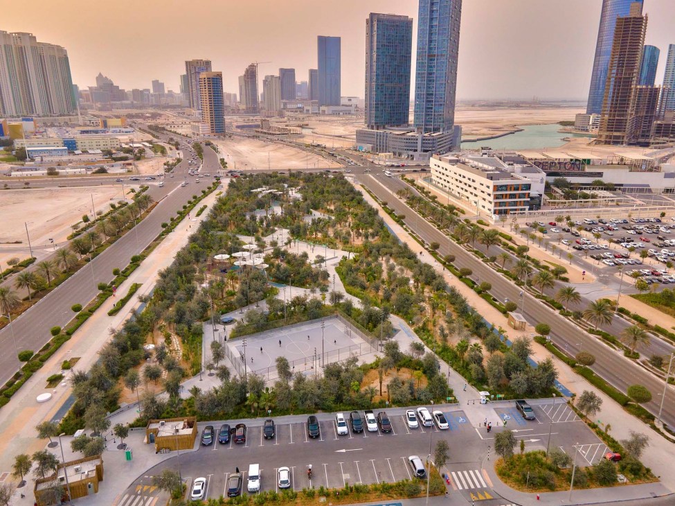 Aerial view of Al Fay Park in Abu Dhabi showing native trees, walking paths, sports courts and no traditional lawns surrounded by desert and high-rise buildings