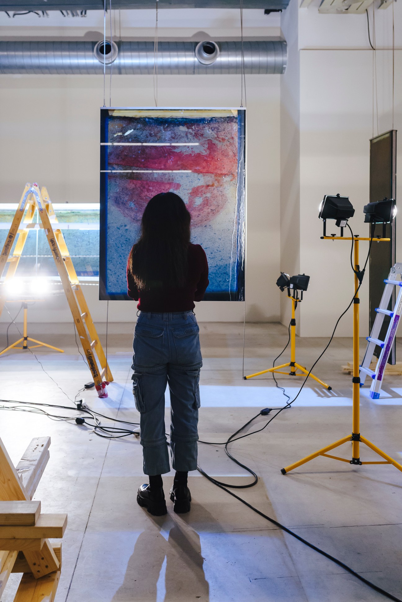 Person with long dark hair stands facing colorful abstract artwork in professional photography setup with lighting equipment