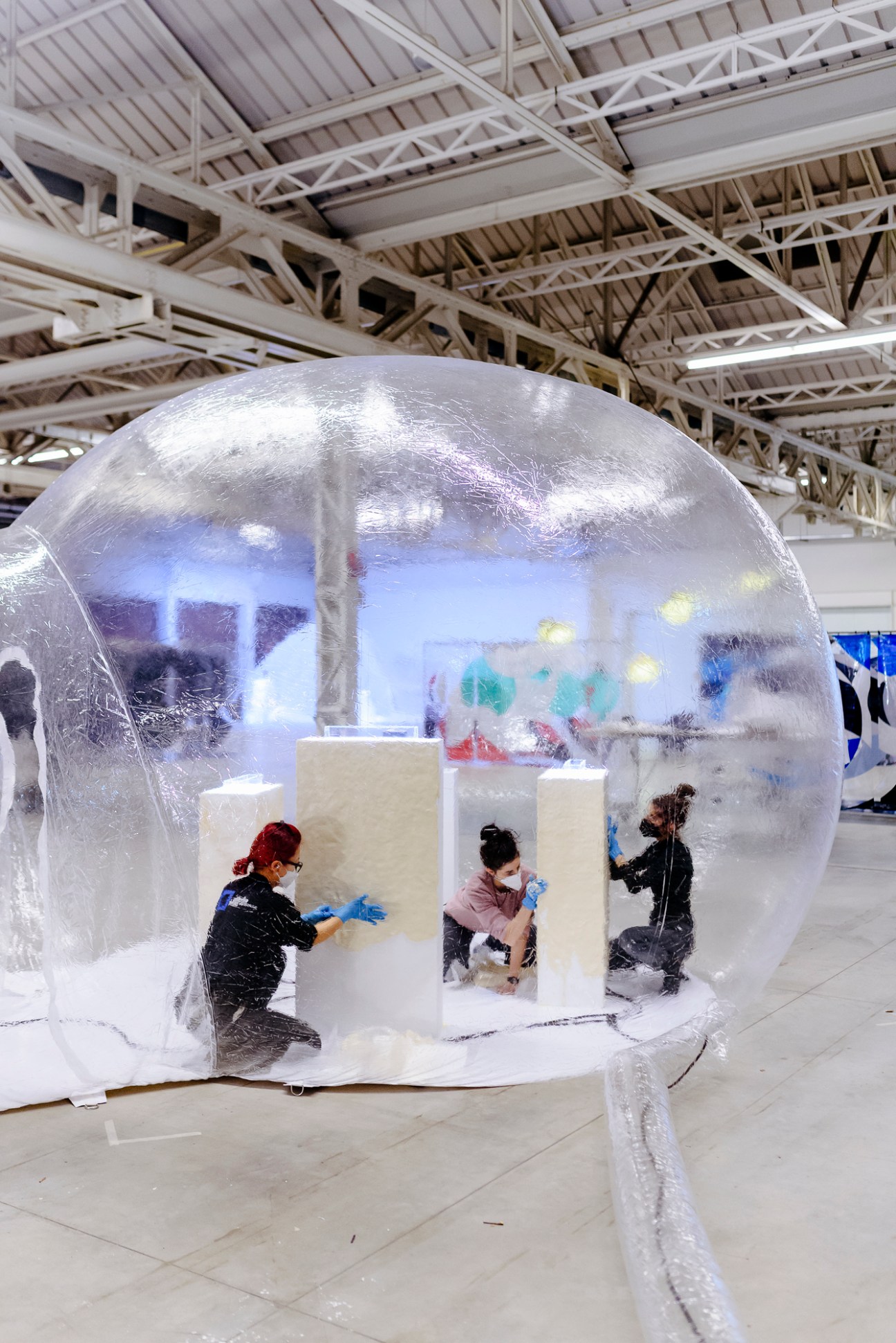 Three people in masks working inside a large transparent inflatable bubble installation in a warehouse space