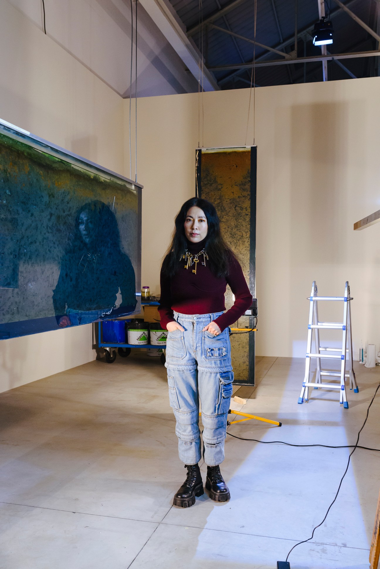 Anicka Yi stands in an art studio wearing dark clothing and a gold necklace, with paintings and equipment visible