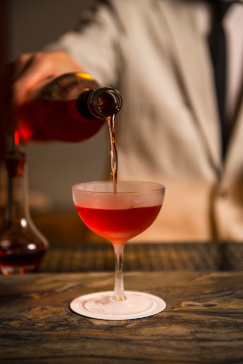 Red cocktail being poured from bottle into coupe glass on wooden bar surface
