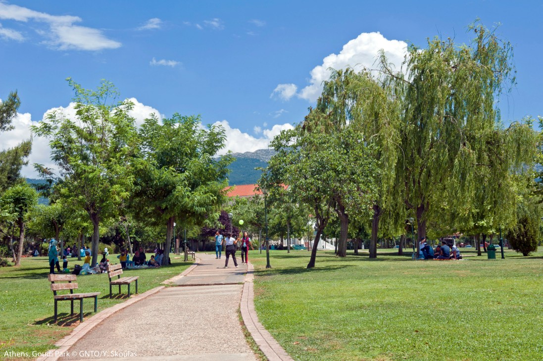 Goudi Park in Athens with tree-lined pathways, green lawns, benches, and visitors enjoying the outdoor space under blue skies.