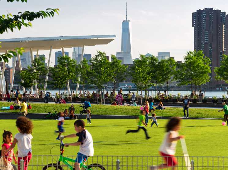 Children play on a grass oval in a waterfront park with One World Trade Center visible in the background.