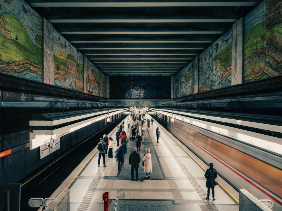 Vienna U-Bahn station platform with passengers waiting between two tracks and decorative murals on the walls.