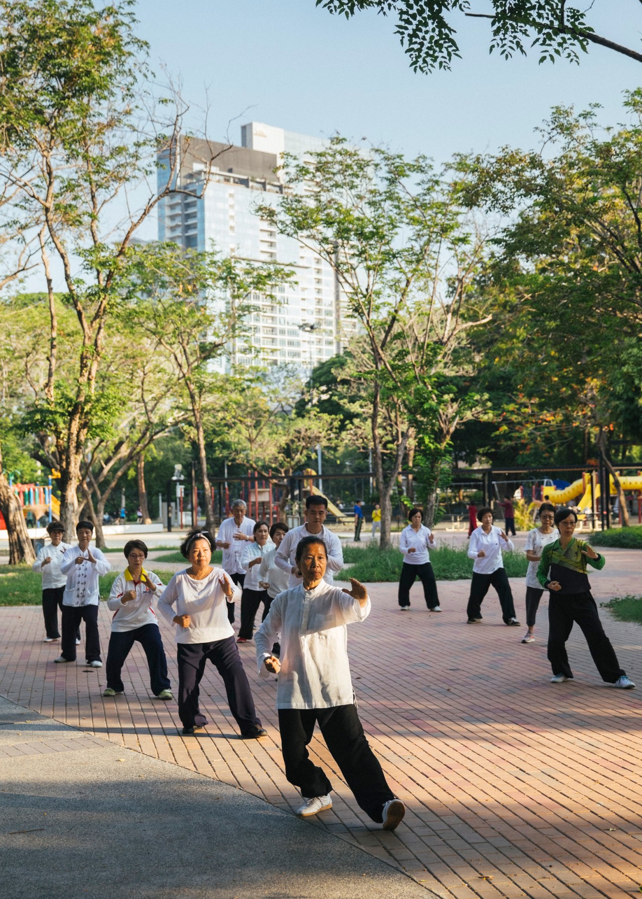 People practicing martial arts in a Bangkok park with trees and a residential building in the background.