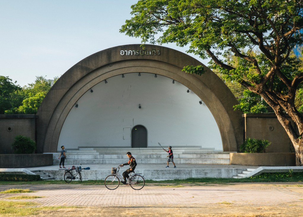Children and young people playing and exercising in front of a large concrete arch structure in Bangkok.