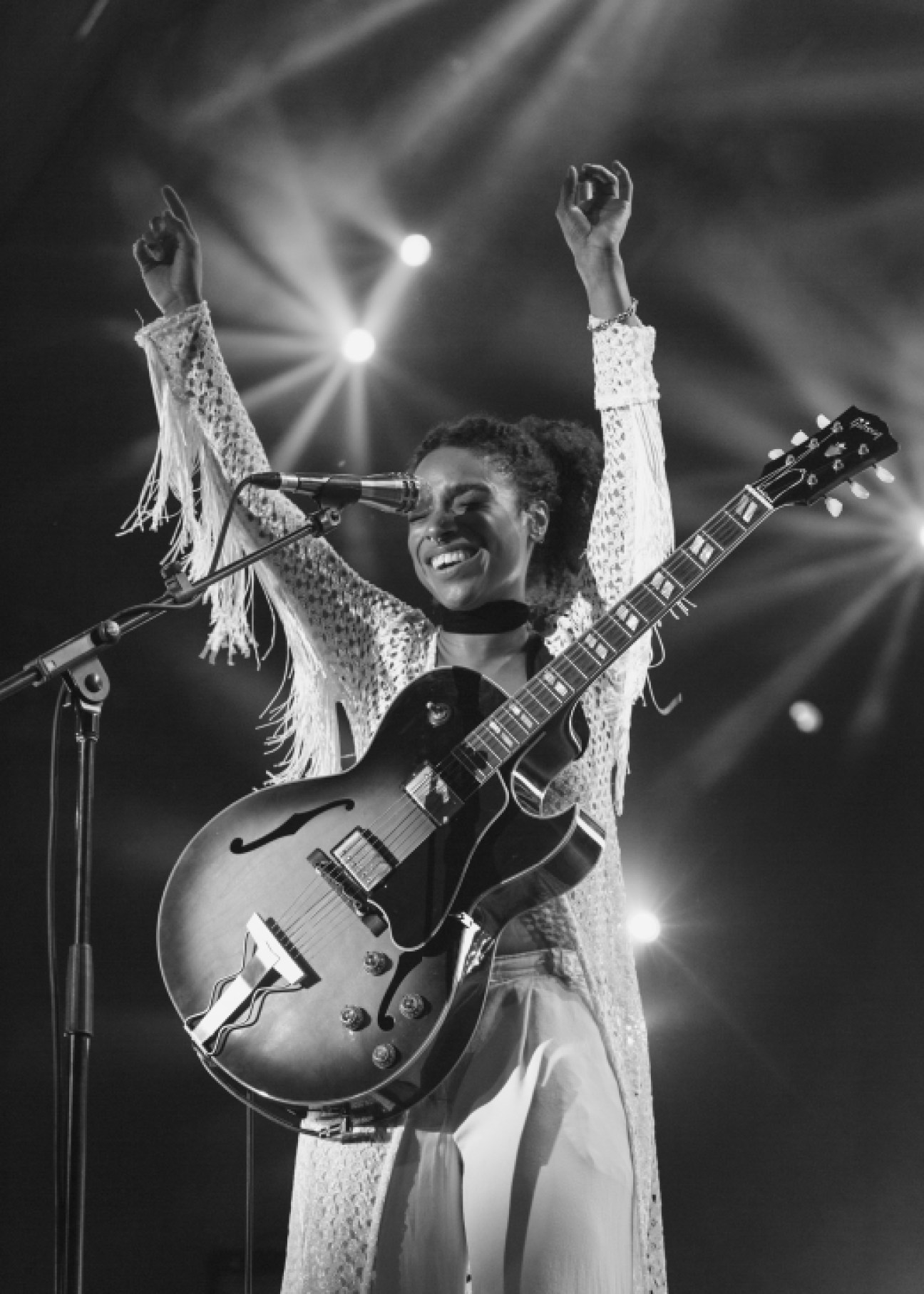 Lianne La Havas performing on stage with guitar, arms raised, wearing a sparkly fringed jacket.