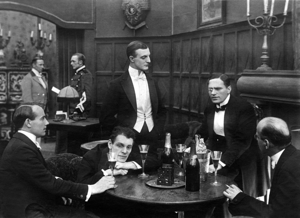 Black and white photograph of men in formal evening wear socializing in an ornate Victorian-era club interior