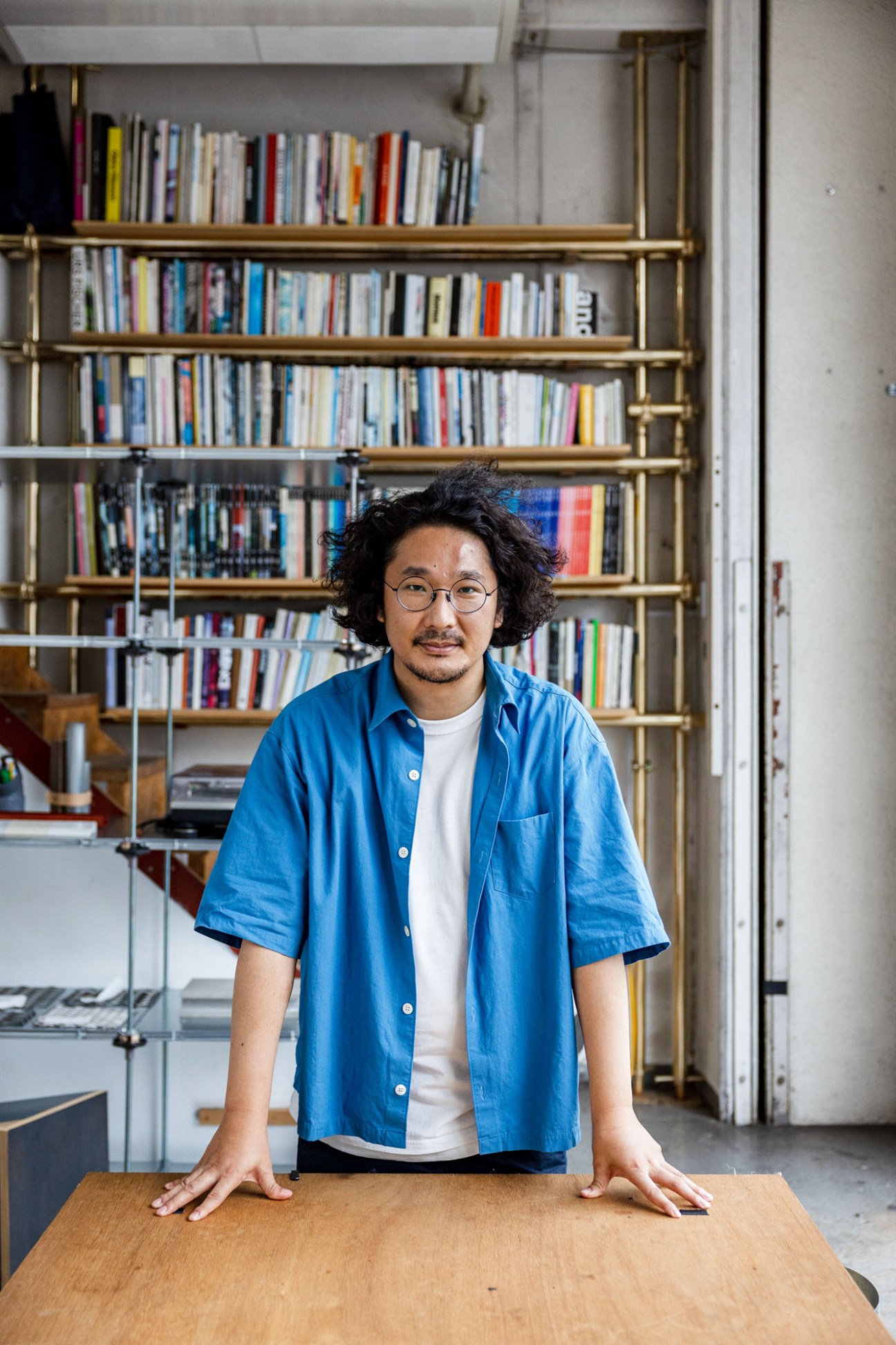 Man with curly hair and glasses wearing blue shirt standing behind wooden table in office with bookshelves