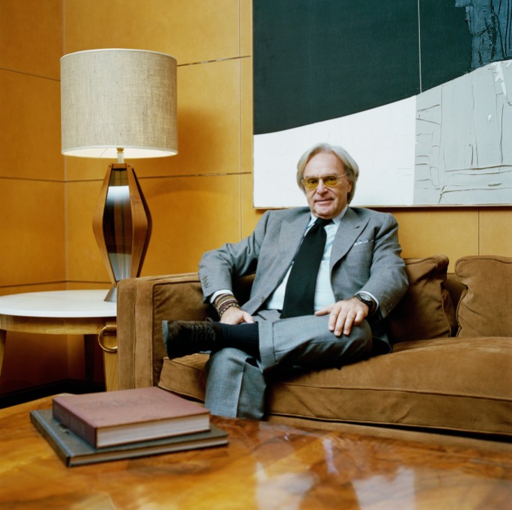 Man in grey suit and yellow glasses sitting on brown leather sofa in mid-century modern interior with yellow walls and table lamp.