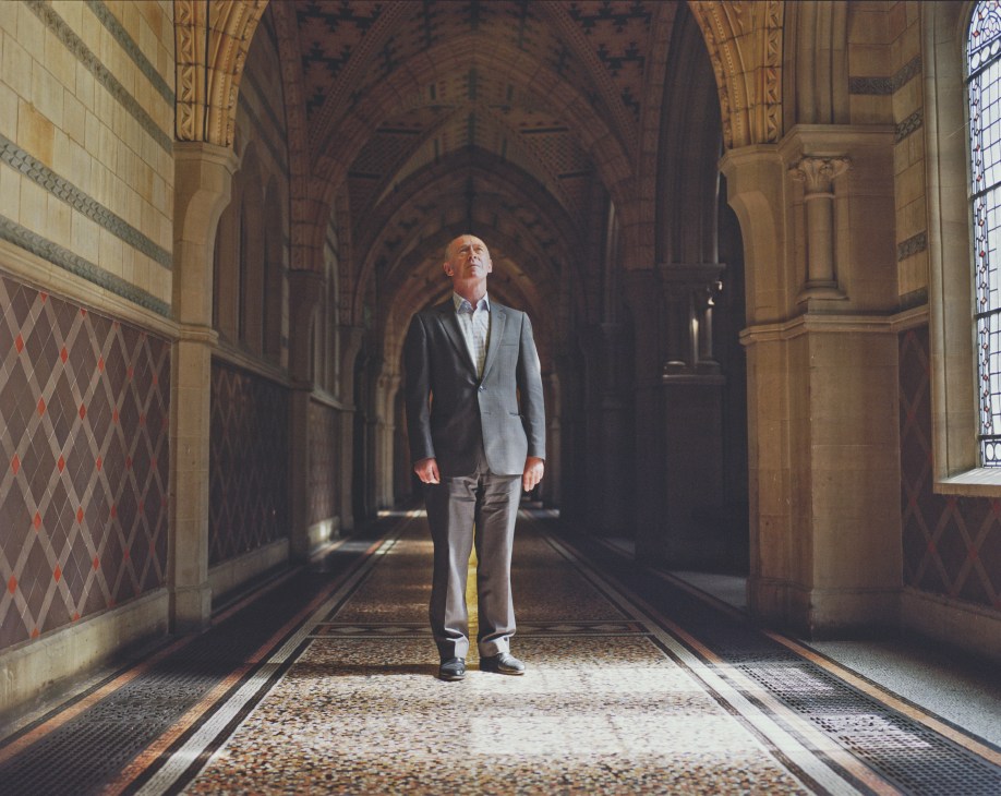 Man in blazer standing in ornate gothic hallway with arched ceilings and decorated walls.
