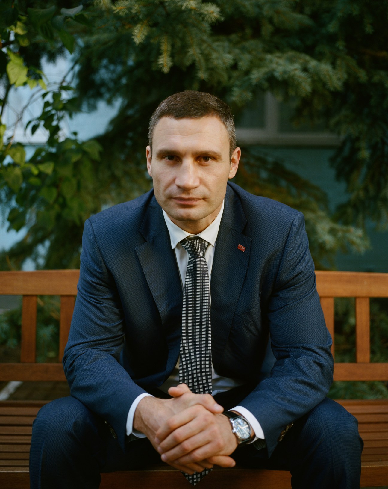 Man in dark suit and patterned tie sitting on wooden bench with tree foliage in background.
