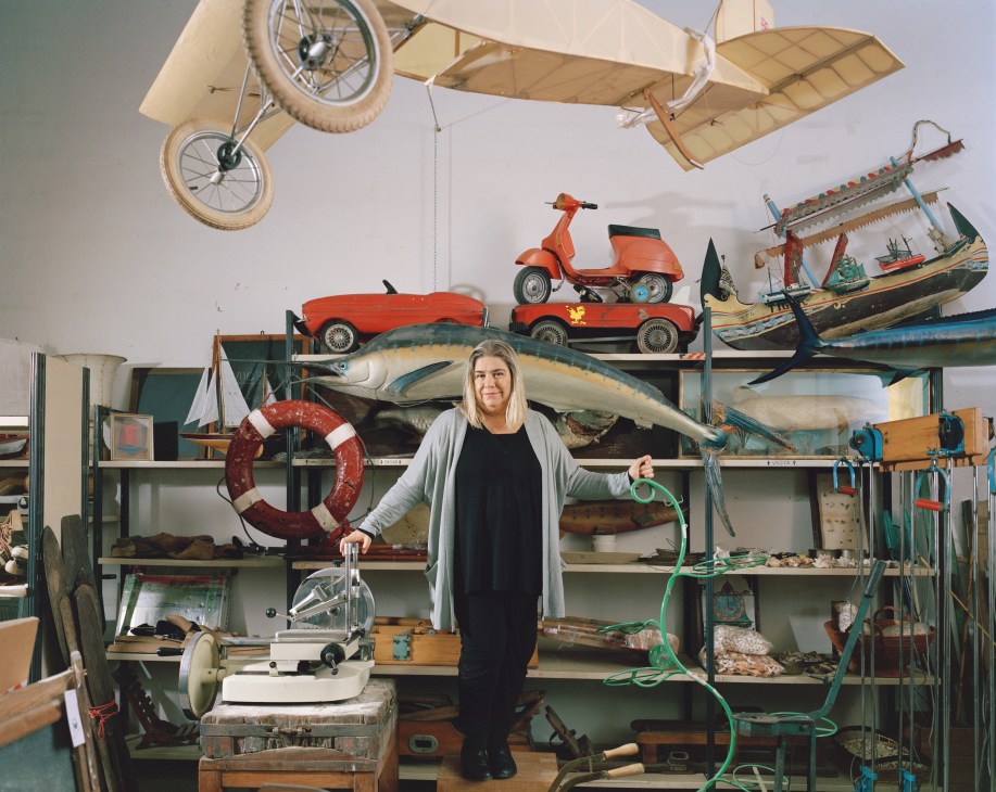 Cristina Calori stands in a storage room surrounded by vintage toys, fishing equipment, and maritime memorabilia.