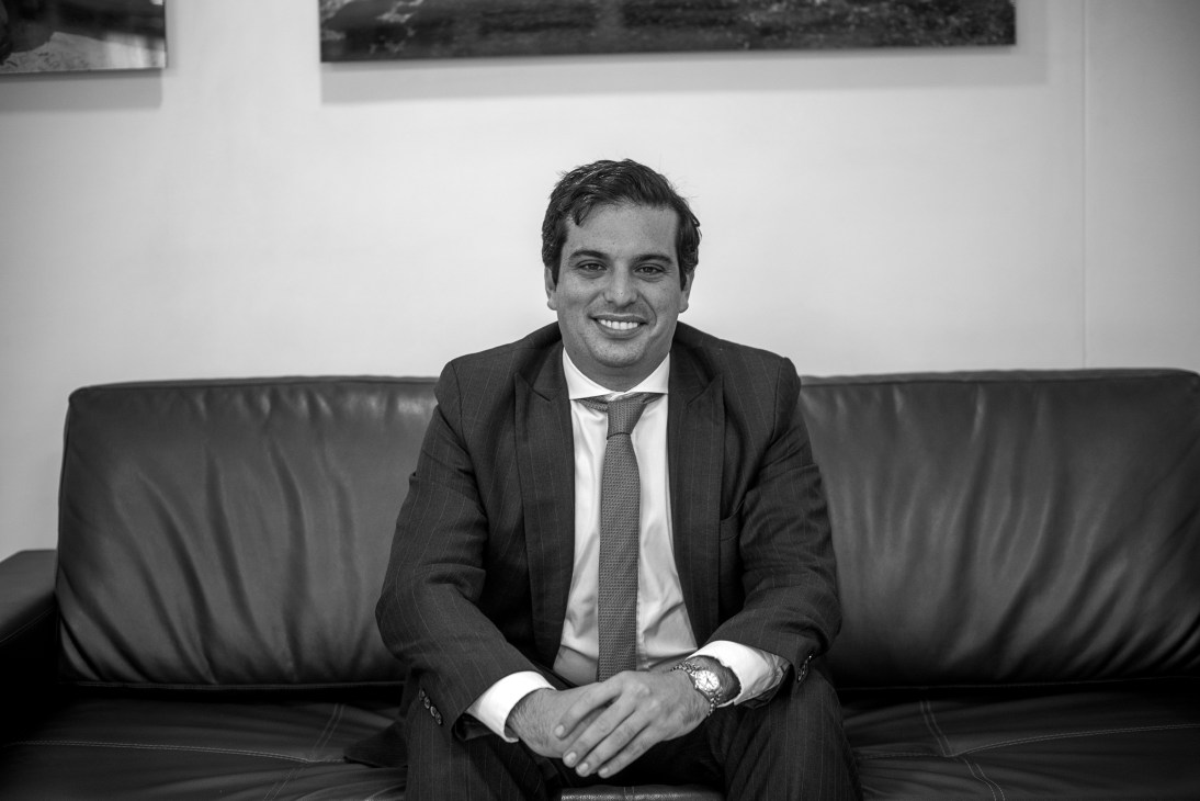 Man in dark pinstriped suit and gray tie smiling while seated on black leather sofa