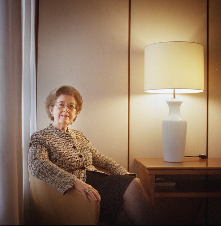 Woman in beige bouclé jacket seated by a window with a lamp, wearing glasses and rings.