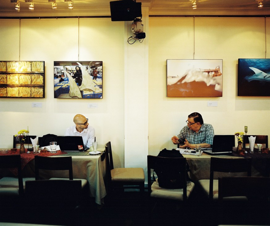 Two journalists work at desks in a modern office space with framed photographs displayed on cream-colored walls.