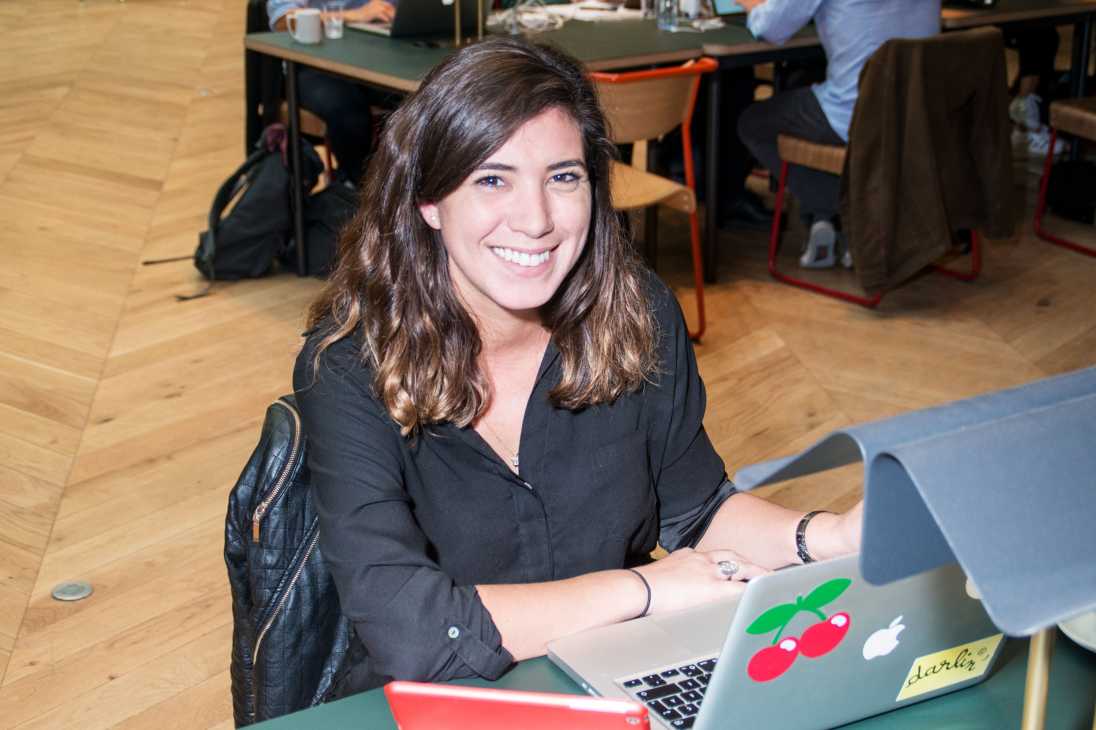 Woman smiling at laptop in co-working space with wooden floor and other workers at tables behind her.