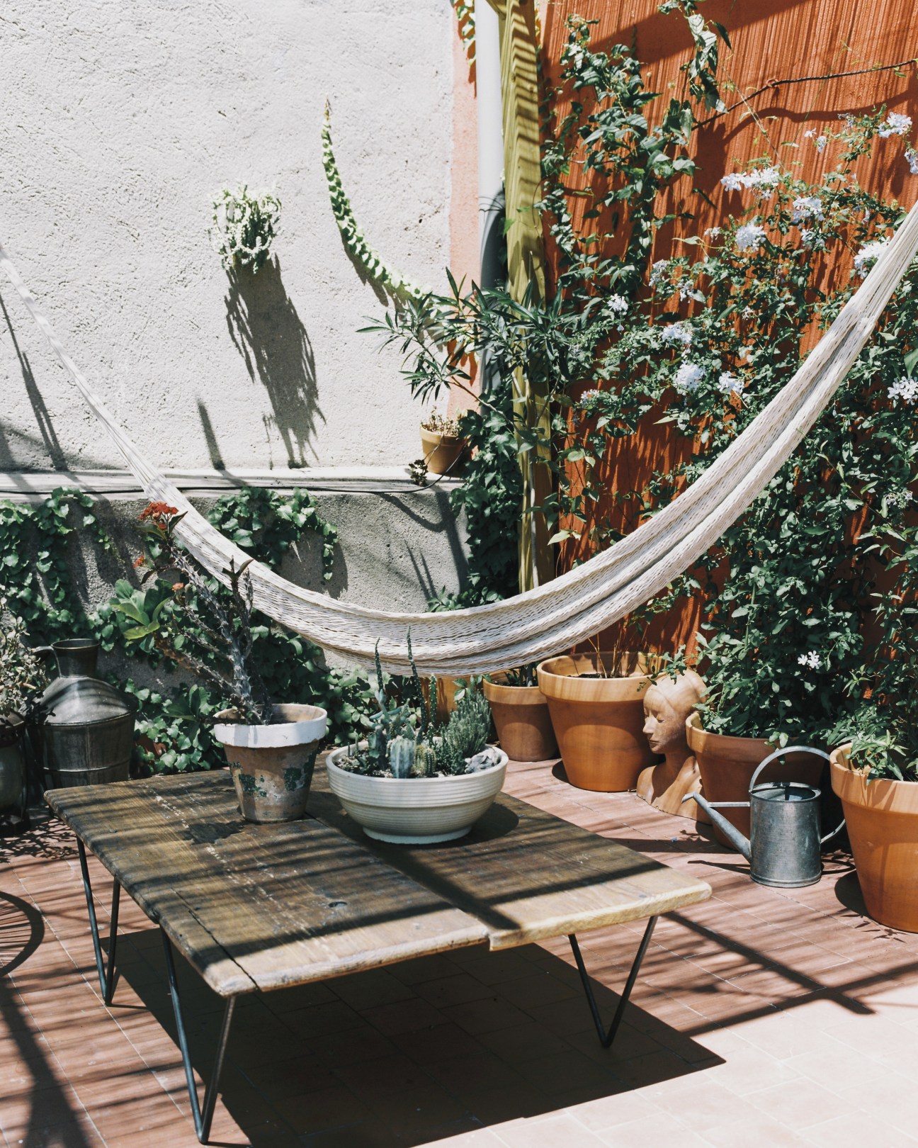 Sun-drenched terrace with potted plants, wooden bench, and hammock against white walls.
