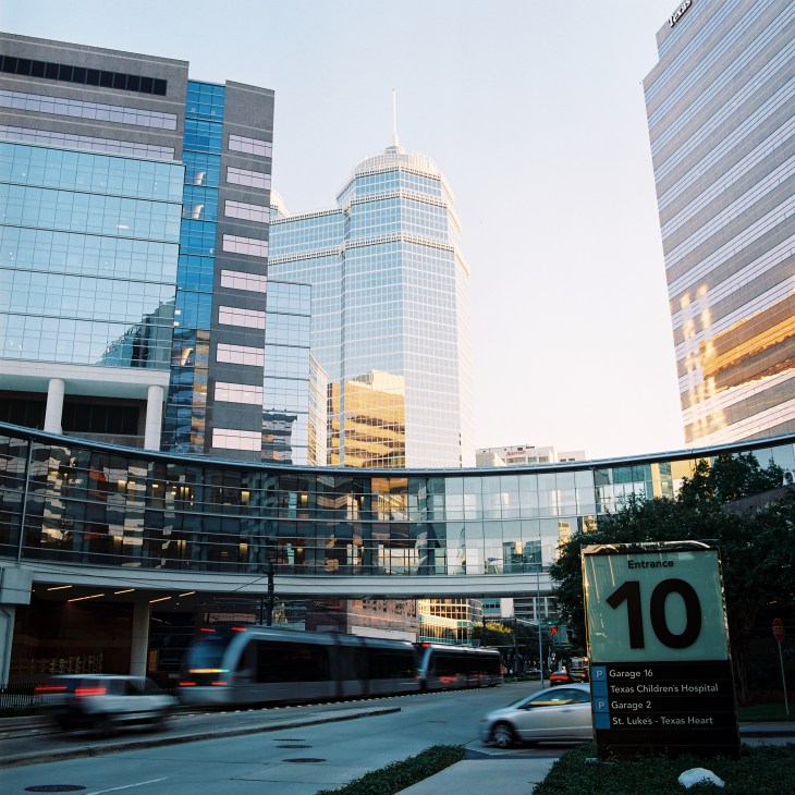 Modern medical buildings and skyscrapers with a metro train passing below an aerial walkway at Texas Medical Center in Houston.