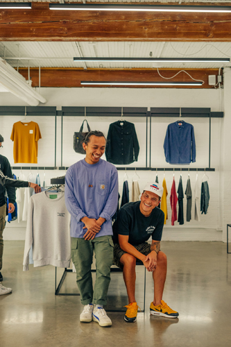 Two men pose inside a retail clothing store with shelves of folded garments and hanging clothes visible behind them.