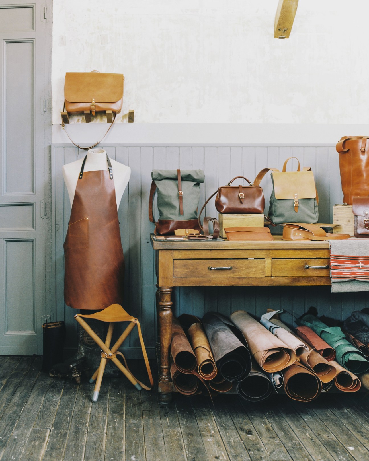 Leather bags, aprons, and rolled hides displayed in Oficio Studio's workshop with wooden workbench and tools.