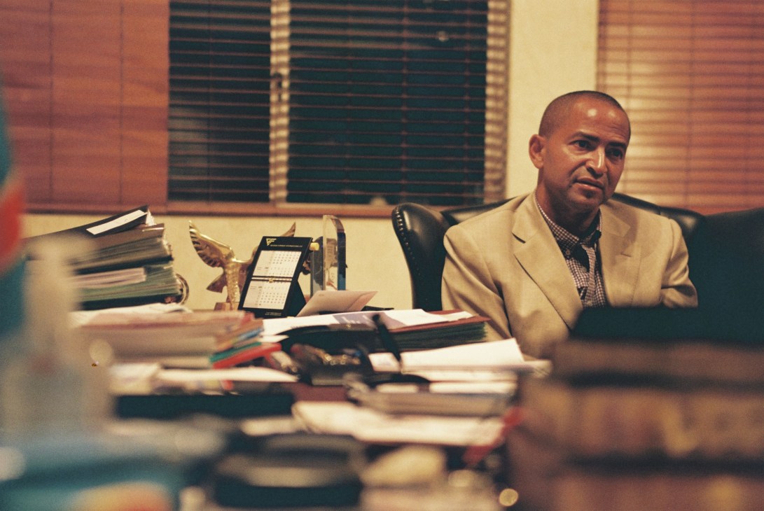 Man in beige suit seated at desk with papers and books in office setting.