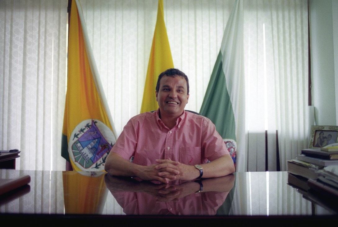 Moïse Katumbi, Governor of Katanga in the Democratic Republic of the Congo, sits at his desk wearing a pink shirt, smiling at the camera with flags behind him.
