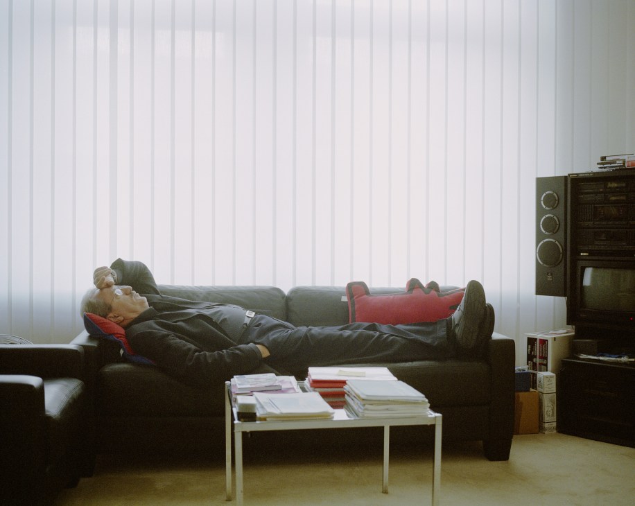 Man in dark suit resting on a black sofa with red pillows in a minimalist office interior.