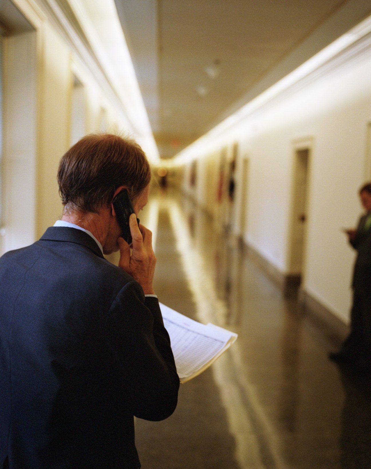 Man in dark suit talking on mobile phone while holding papers in a bright corridor.