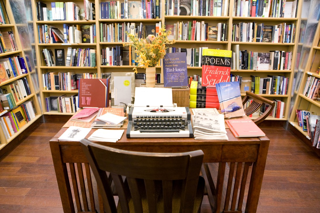 Wooden desk with typewriter, papers, and flowers in front of tall wooden bookshelves filled with books.