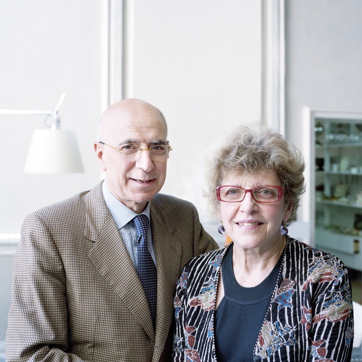 Two people standing together in a bright, modern interior with shelving visible in the background.