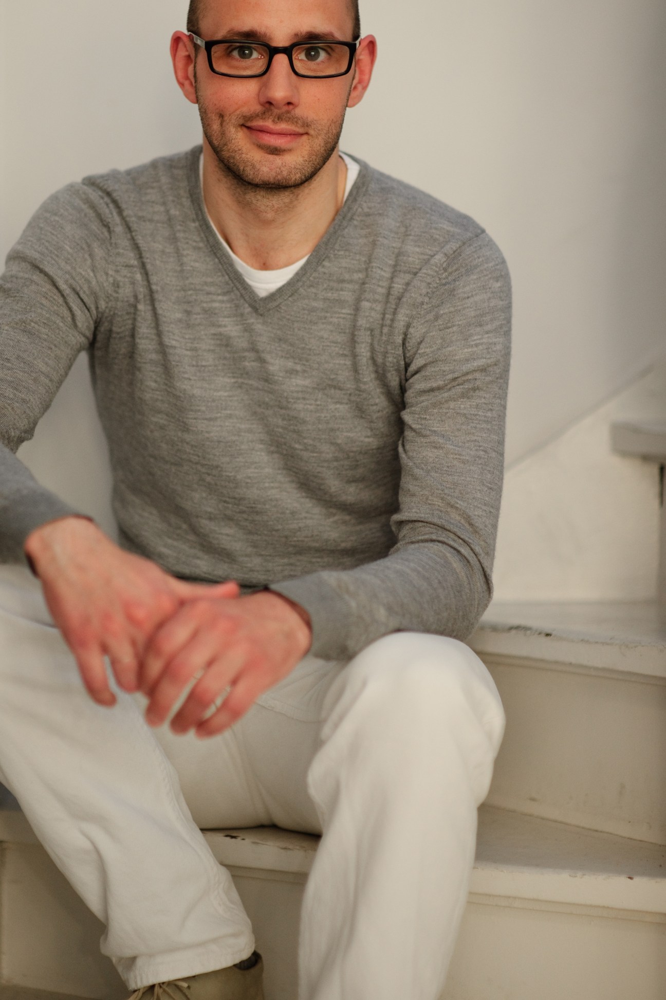 Man wearing glasses and grey V-neck sweater with white pants sitting indoors.