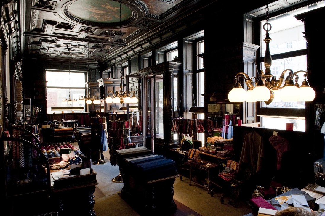 Wilhelm Jungmann & Neffe tailoring shop interior in Vienna with ornate ceiling, brass chandeliers, and fabric displays.