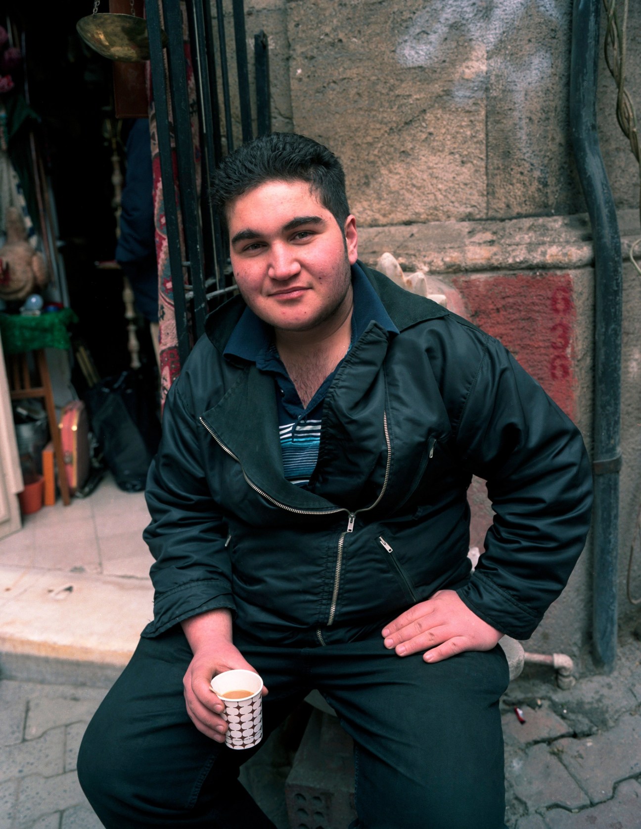 Young man in black jacket holding a patterned cup, seated on steps against a weathered stone wall on Turnacibasi Sokak in Istanbul.