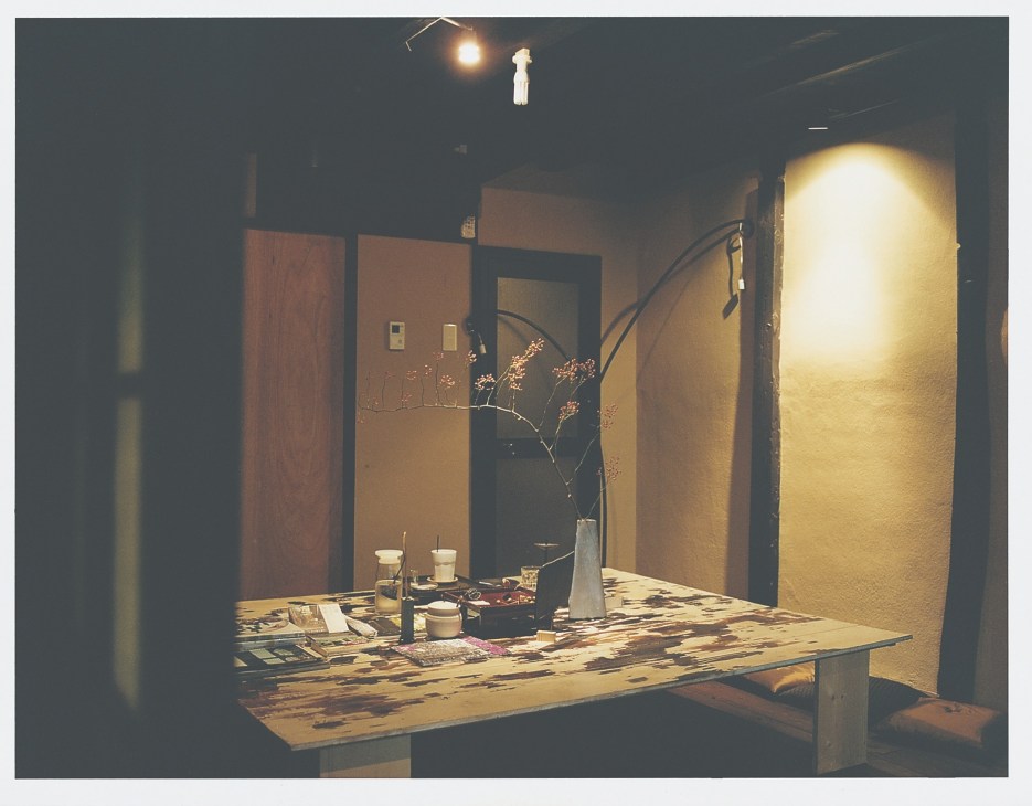 A dimly lit artisan workshop with warm overhead lighting, wooden work table displaying crafts, and dried flowers in a vase.