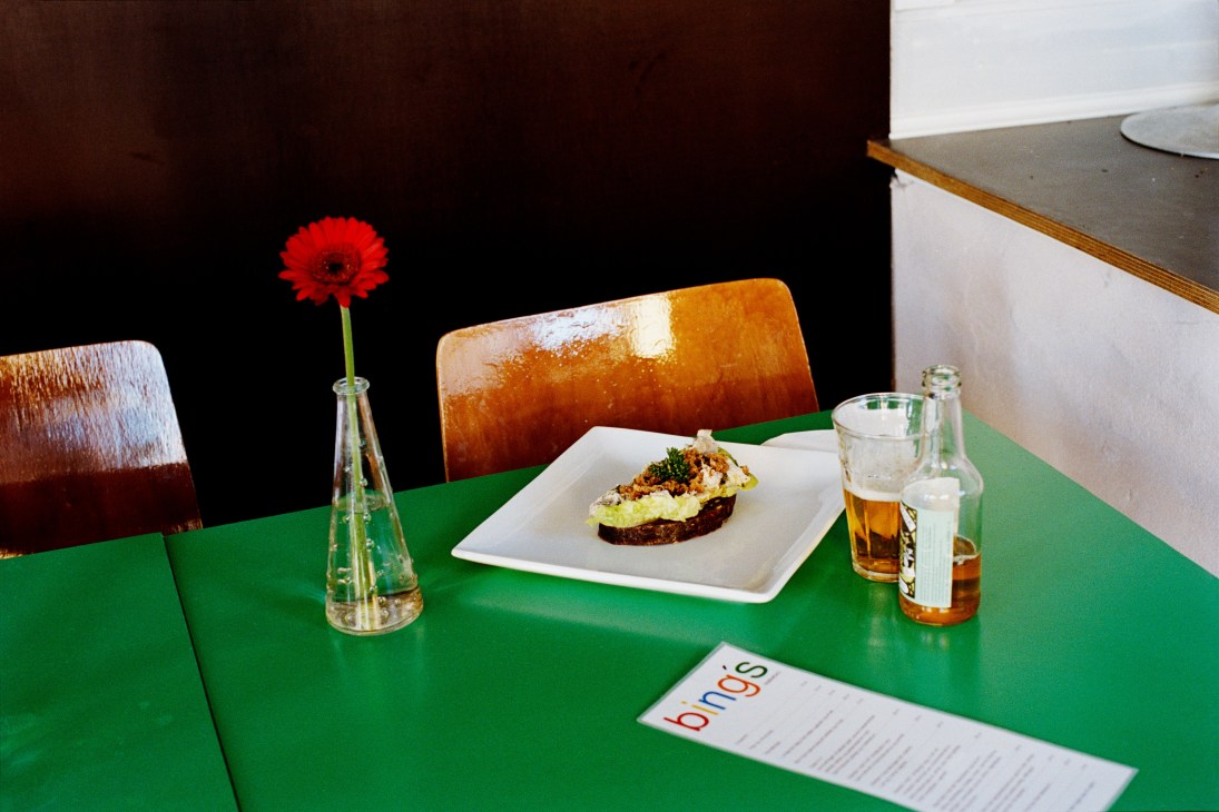 A bright green table set with wooden chairs, a red gerbera flower, plated food, drinks, and a Binos menu.