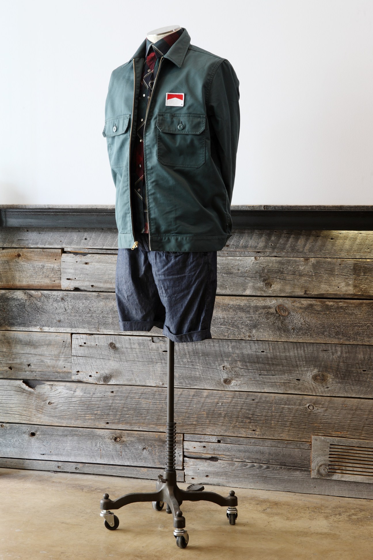 Mannequin displaying a dark green utility jacket with Indonesian flag patch and dark denim shorts against wooden backdrop.