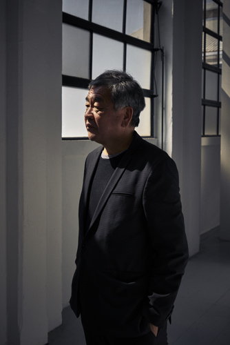 Architect Kengo Kuma stands in a minimalist white corridor with large windows.