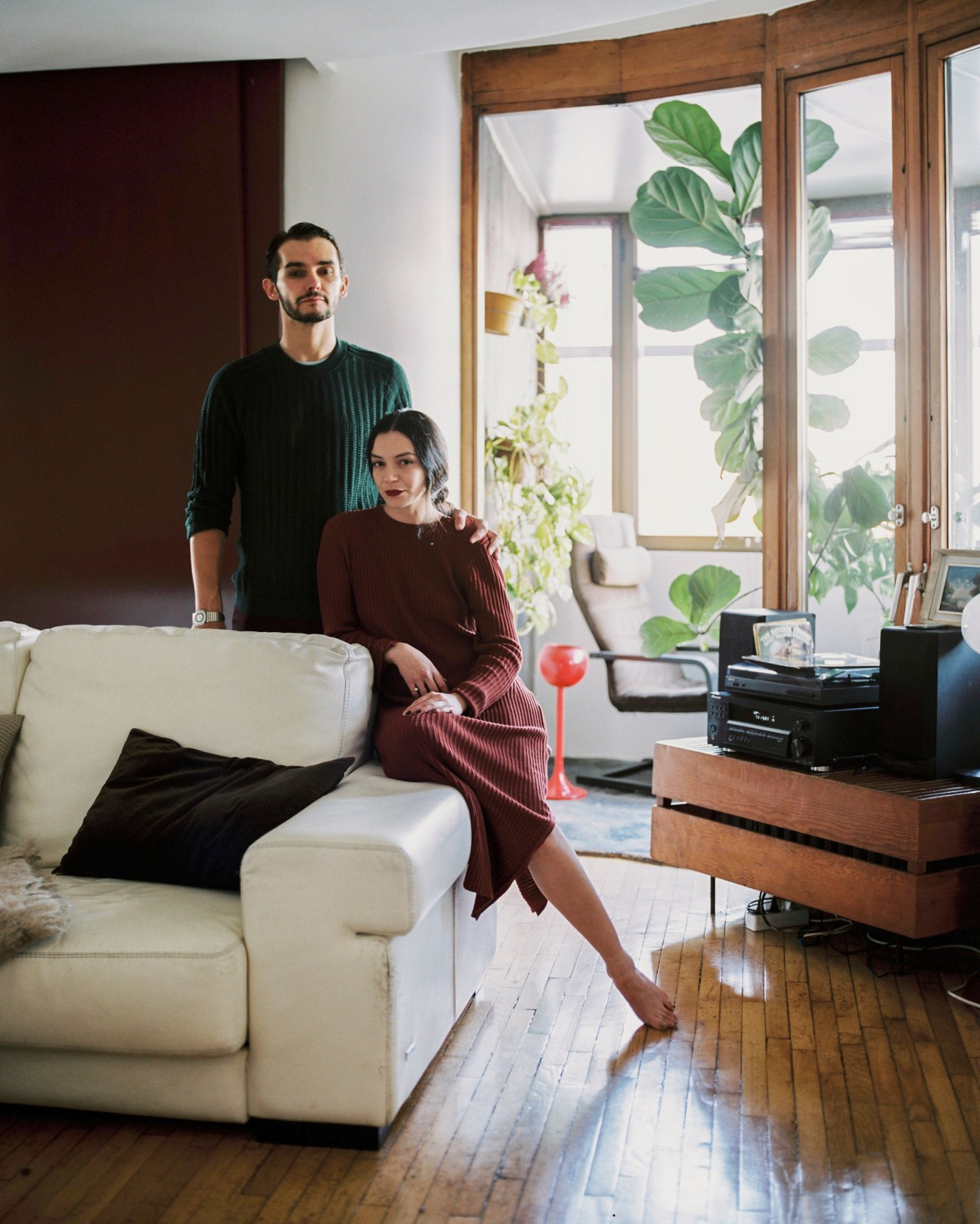 Two residents stand in a modern living room with a white sofa, wooden furniture, and large windows overlooking green plants.