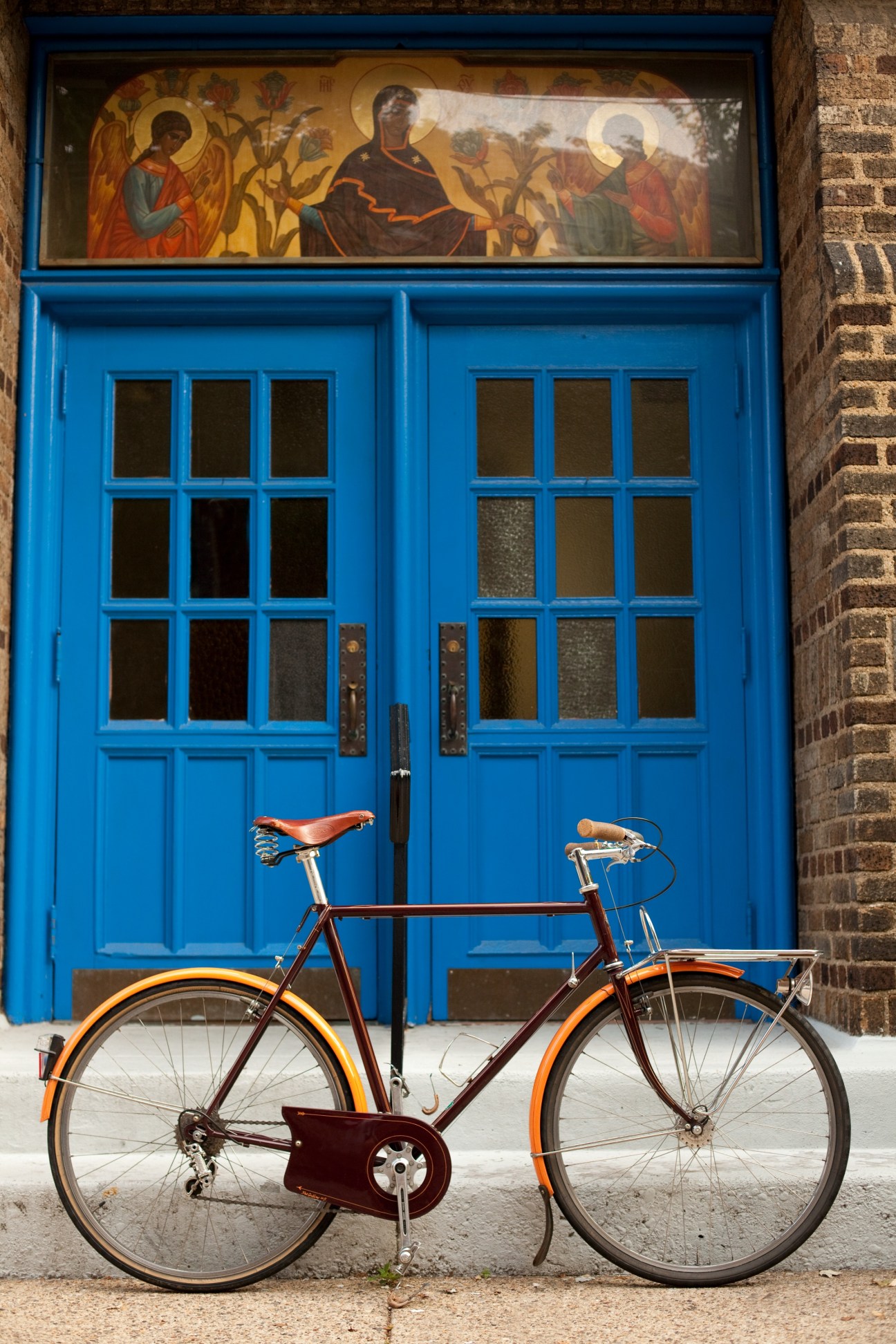 A burgundy and orange Bilenky Porteur bicycle leaning against a bright blue door with religious iconography above it.