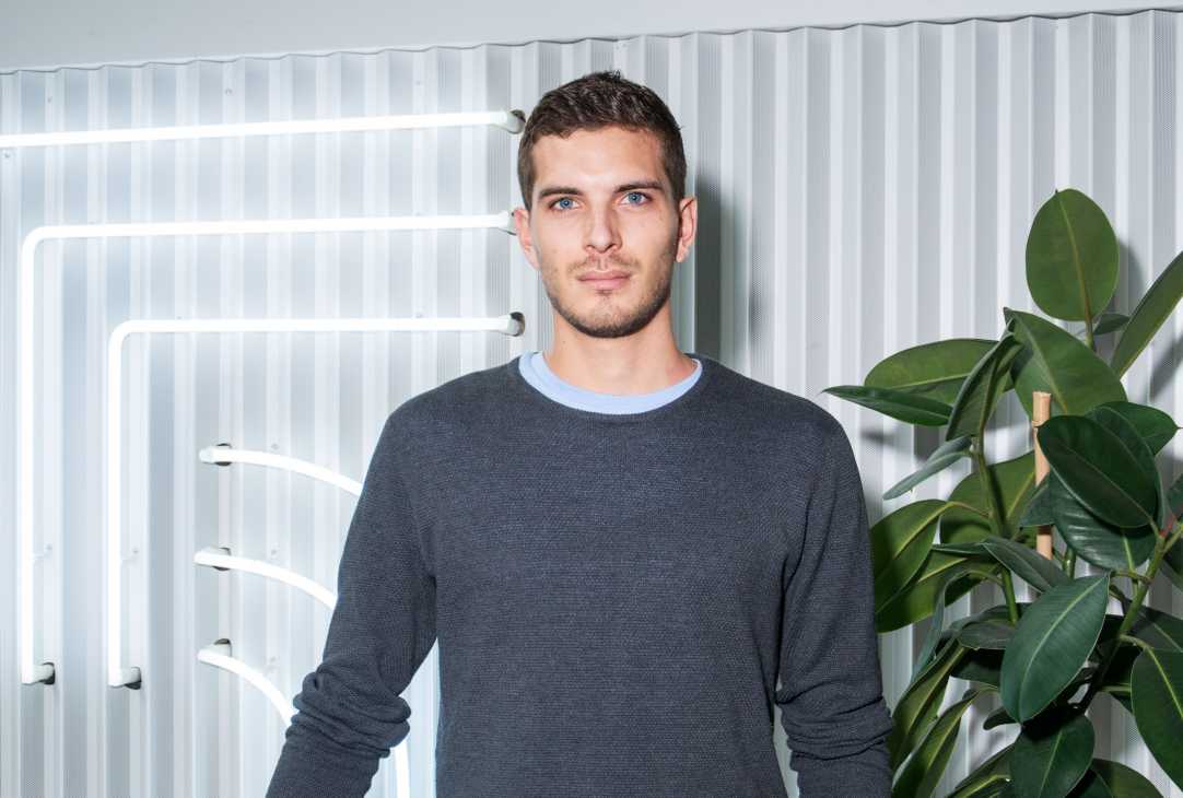 Man in dark sweater standing against corrugated metal wall with neon lighting and potted plant