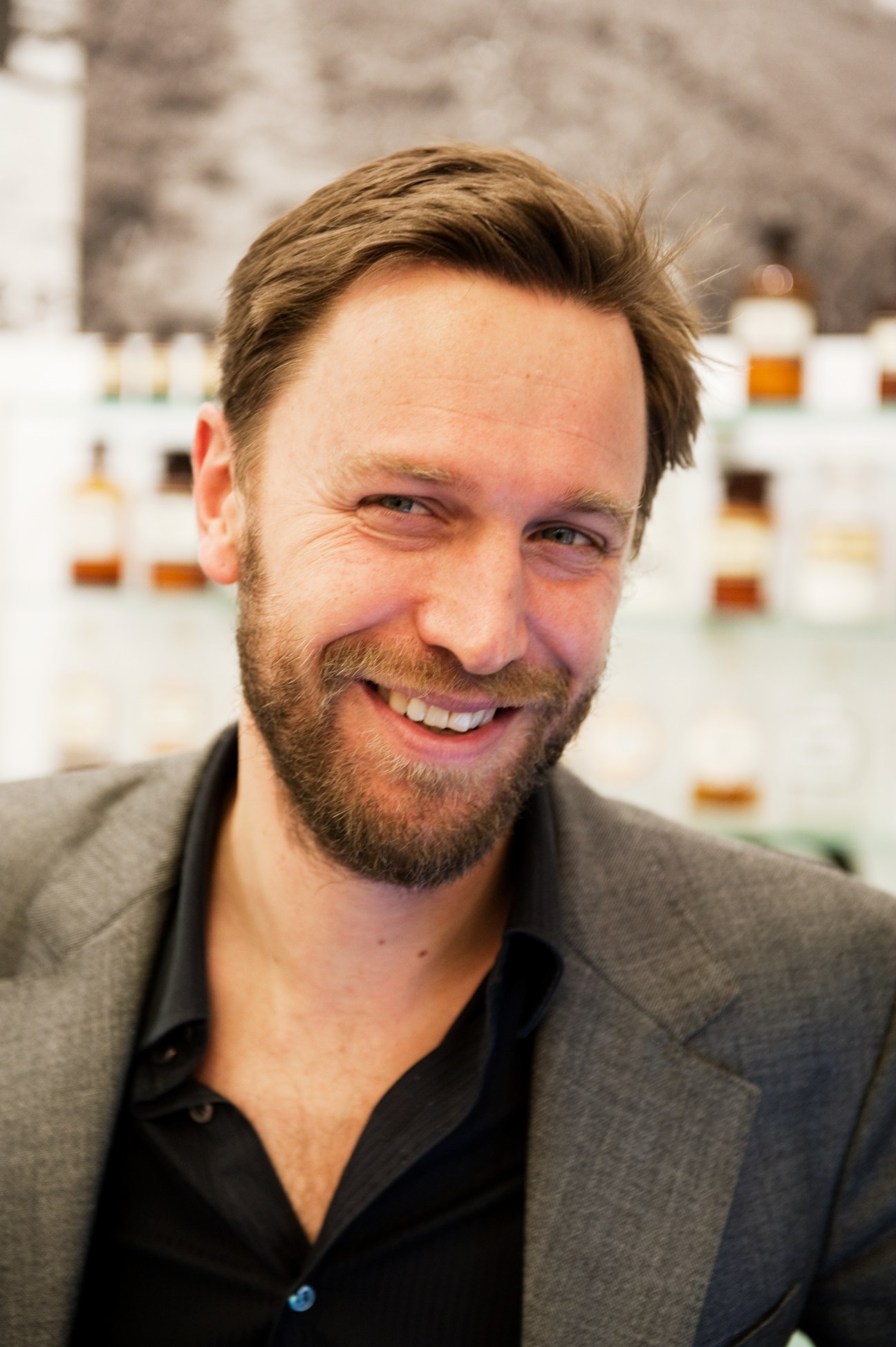 Portrait of a bearded man in a grey blazer and black shirt smiling at the camera.