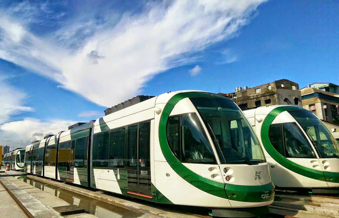 White and green Kaohsiung Light Rail trams lined up on tracks with buildings in background.
