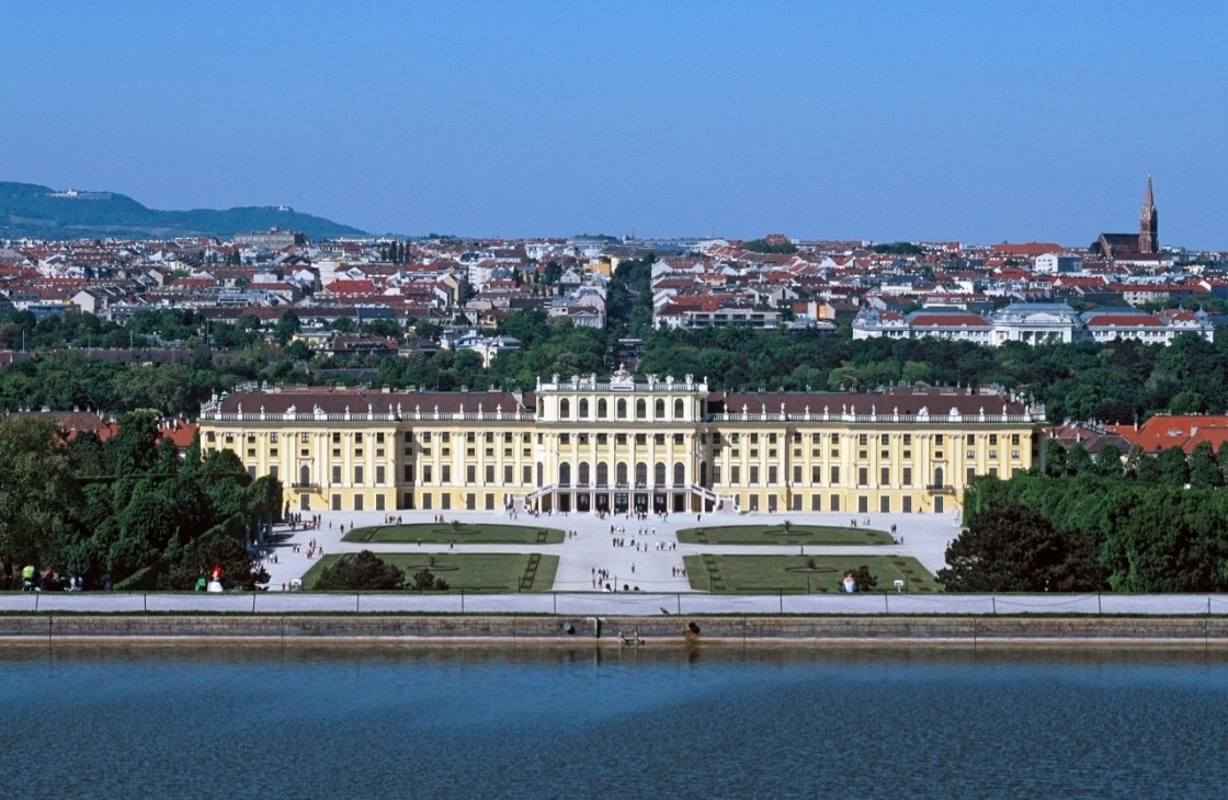 Schönbrunn Palace's yellow facade overlooks formal gardens and the Danube River with Vienna's cityscape beyond.