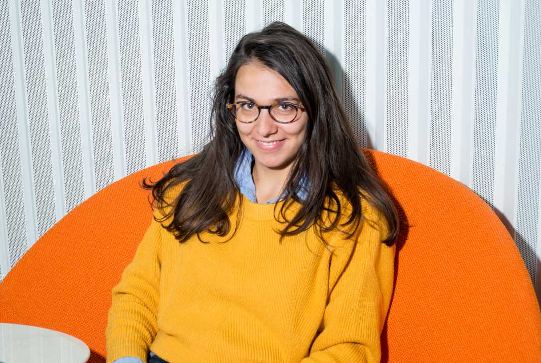 Woman with black hair and glasses wearing a yellow sweater seated on an orange chair.