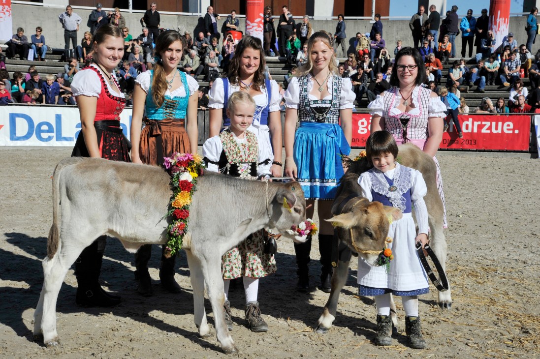 Young women and children in traditional Alpine clothing pose with dairy cattle at an agricultural fair.